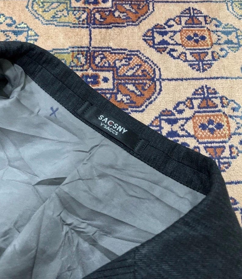 May include: Close-up of a dark gray garment with a black label that reads "SACSNY Y'SACCS". The interior lining is a light gray color. The garment is on a patterned beige and brown surface.