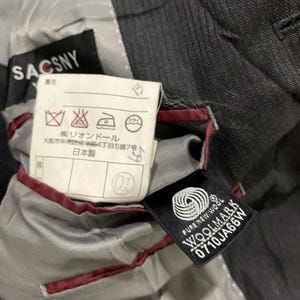 May include: Close-up of the interior of a dark gray suit jacket. The lining is light gray with red trim. Tags include "SACSNY", "PURE NEW WOOL WOOLMARK", and Japanese text with care symbols.