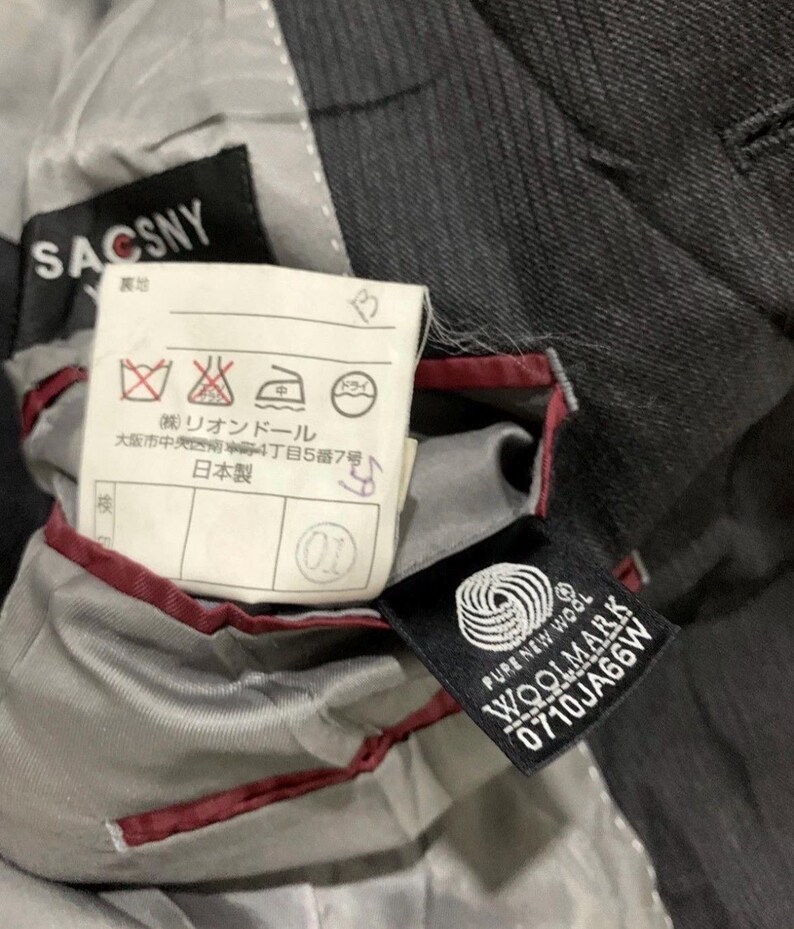 May include: Close-up of the interior of a dark gray suit jacket. The lining is light gray with red trim. Tags include "SACSNY", "PURE NEW WOOL WOOLMARK", and Japanese text with care symbols.