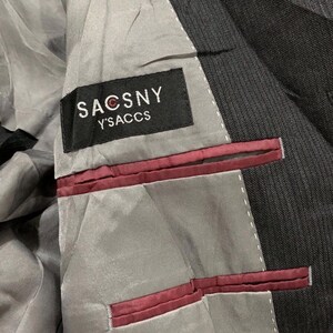 May include: Close-up of the interior of a dark gray suit jacket. The lining is light gray with burgundy accents. A black label reads "SACSNY Y'SACCS" in white lettering.