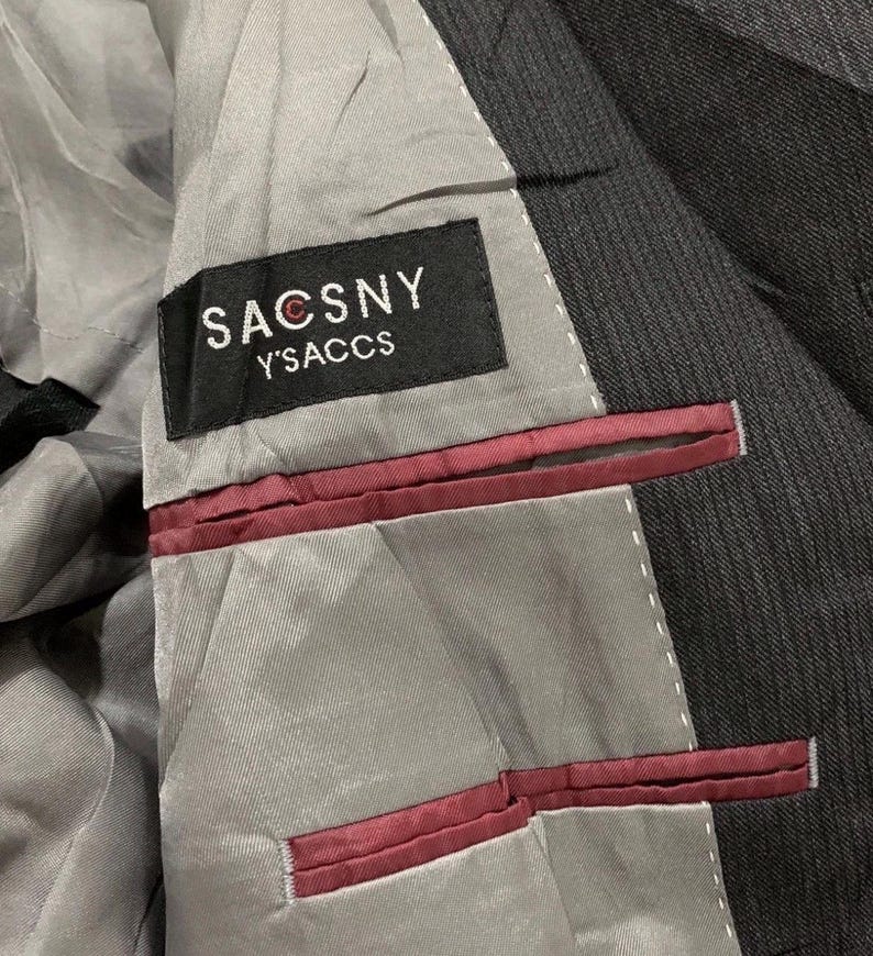 May include: Close-up of the interior of a dark gray suit jacket. The lining is light gray with burgundy accents. A black label reads "SACSNY Y'SACCS" in white lettering.