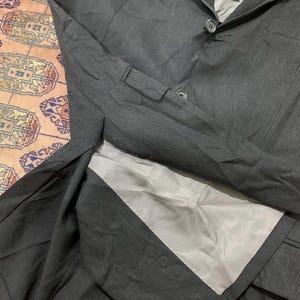 May include: A dark gray blazer with a two-button closure and a notched lapel. The blazer has a textured fabric and a light gray lining. The image shows the blazer laid flat, with the interior visible.