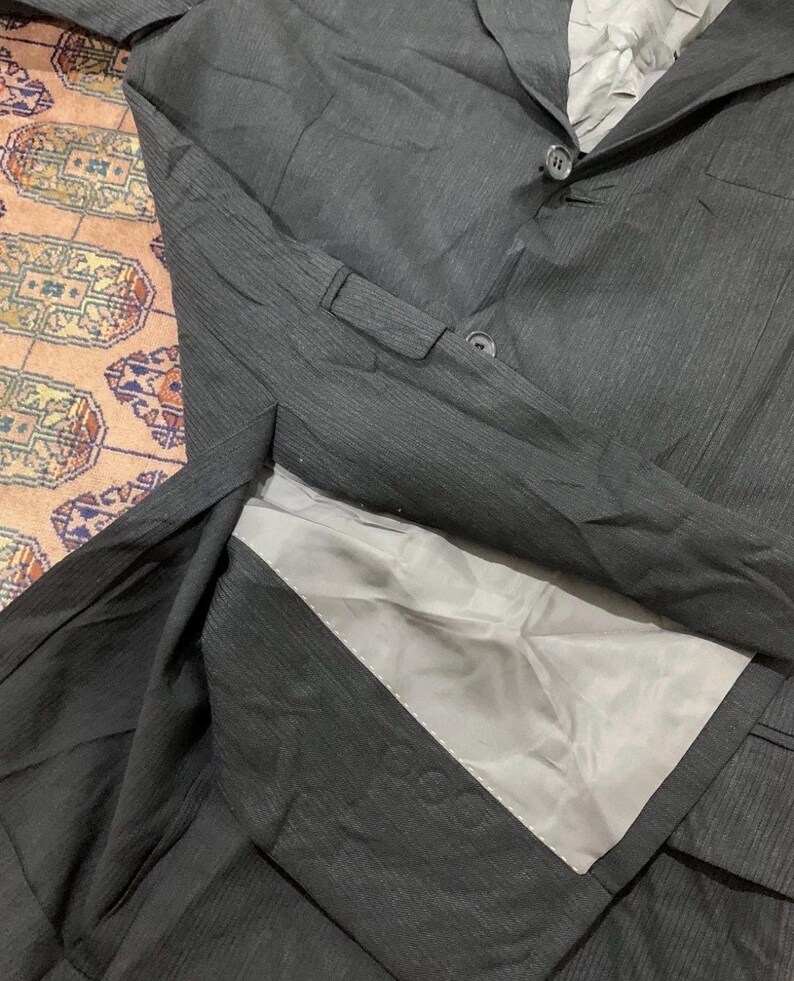 May include: A dark gray blazer with a two-button closure and a notched lapel. The blazer has a textured fabric and a light gray lining. The image shows the blazer laid flat, with the interior visible.