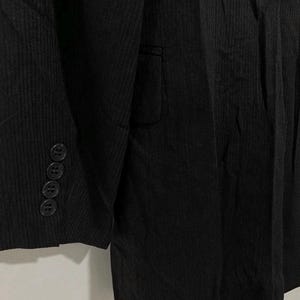 May include: Close-up of a black blazer with a textured, ribbed fabric. The blazer features a notched lapel, a single-breasted design, and four buttons on the sleeve cuff. The fabric has vertical lines, giving it a subtle striped appearance.