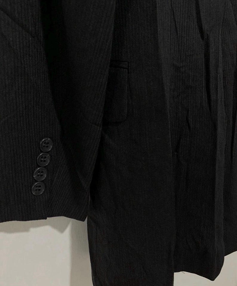May include: Close-up of a black blazer with a textured, ribbed fabric. The blazer features a notched lapel, a single-breasted design, and four buttons on the sleeve cuff. The fabric has vertical lines, giving it a subtle striped appearance.