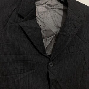 May include: A black blazer with a subtle pinstripe pattern. The jacket features a notched lapel, two front buttons, and a chest pocket. The interior lining is a light gray color. The blazer is hanging on a black hanger.