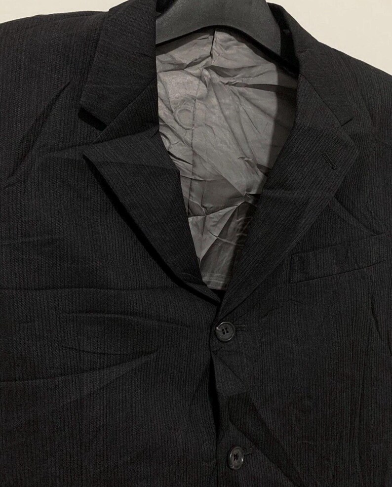 May include: A black blazer with a subtle pinstripe pattern. The jacket features a notched lapel, two front buttons, and a chest pocket. The interior lining is a light gray color. The blazer is hanging on a black hanger.