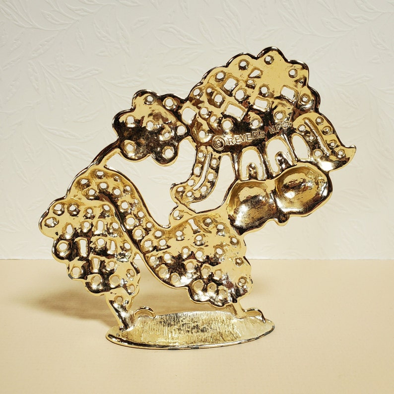 Op de afbeelding: Gouden metalen decoratieve standaard met een boomontwerp. De standaard heeft een ronde basis en is gemaakt van ingewikkeld, opengewerkt metaal. De tekst "REVERE WARE" is zichtbaar op de standaard.