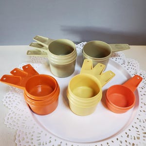 May include: Set of six vintage measuring cups in orange and beige. The cups are made of plastic and have a nested design. The cups are arranged on a white lace doily.