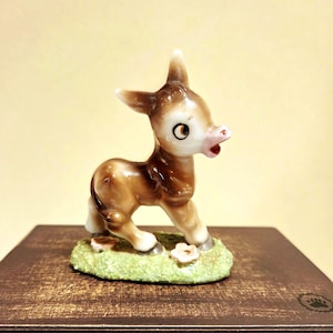May include: A ceramic figurine of a donkey with brown and white coloring. The donkey has a surprised expression with its mouth open. It stands on a small patch of green grass with a white flower. The figurine is a decorative item.