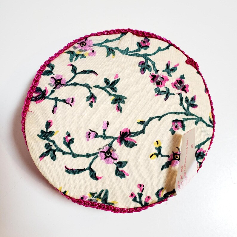 May include: A circular fabric coaster featuring a cream background adorned with a pink and green floral design. The edge is trimmed with a pink, ruffled trim. A small tag is attached to the coaster.