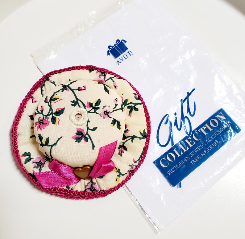 May include: A hat-shaped floral pin cushion with a pink bow and trim. The pin cushion is part of the Avon Gift Collection, which includes Victorian sewing accessories and a tape measure.