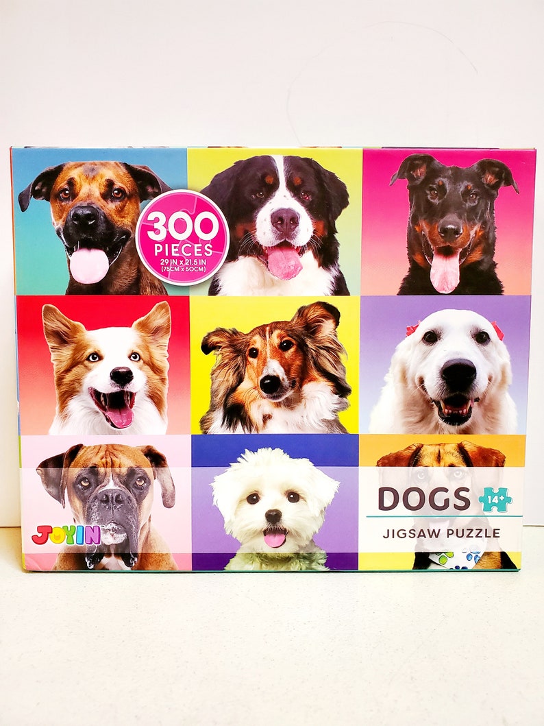 May include: A jigsaw puzzle box featuring nine different dog breeds against colorful backgrounds. The puzzle contains 300 pieces and measures 29 inches by 21.5 inches. The box has the word "DOGS" and "JIGSAW PUZZLE" printed on it.