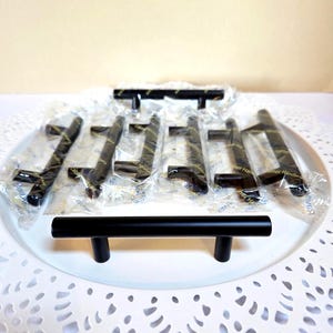 May include: A collection of black cabinet pulls, some individually wrapped, arranged on a white plate. The pulls have a sleek, modern design, with a bar and two vertical posts. The hardware is likely for kitchen or bathroom cabinets.
