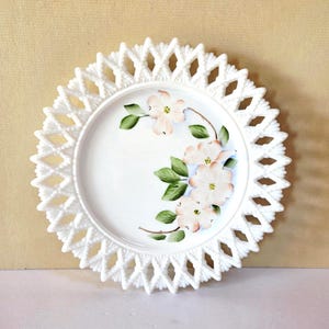 May include: White decorative plate with a pierced, openwork border. The center of the plate features a floral design with pink and white flowers and green leaves. The plate has a diameter of approximately 8 inches.
