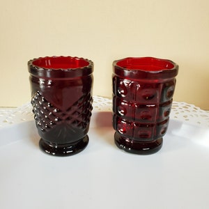 May include: Two small, red glass candle holders with different patterns. The holder on the left has a diamond pattern and the holder on the right has a square pattern.