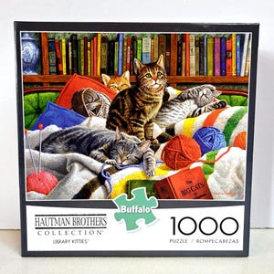 May include: A 1000-piece puzzle featuring a cozy scene of cats lounging on a bed with yarn balls and books. The puzzle box has the text "Library Kitties" and "Buffalo" on it.