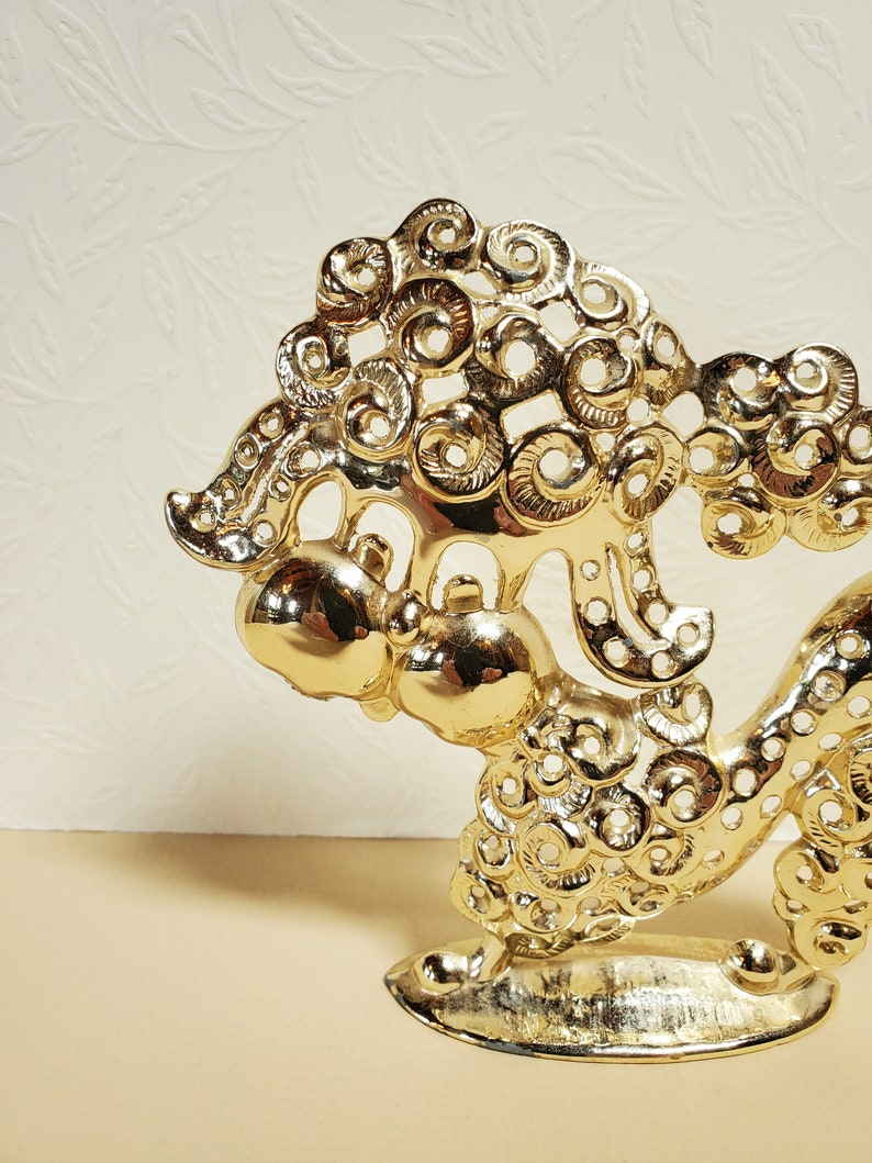 Op de afbeelding: Een gouden metalen beeldje van een gestileerde draak met ingewikkelde details. De draak is opgerold met zijn kop omhoog en zijn staart om zijn lichaam gewikkeld. Het beeldje is gemaakt van massief metaal en heeft een gladde, gepolijste afwerking.