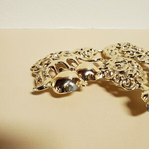 Op de afbeelding: Een gouden metalen broche met een ingewikkeld ontwerp van draaiende patronen en afgeronde vormen. De broche heeft een delicate en sierlijke uitstraling.