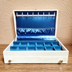 May include: Open white jewelry box with a blue velvet interior and a blue satin lining. The box has multiple compartments for storage and small gold-colored knobs on the front.