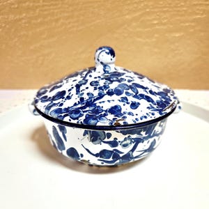 May include: A round enamel bowl with a matching lid, featuring a white base with a blue splatter pattern. The bowl has two small side handles and a knob on the lid. The rim of the bowl and the lid are finished with a dark blue metal edge.