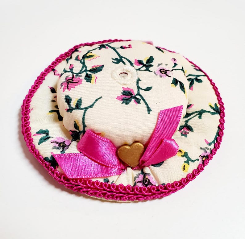 May include: A miniature hat with a floral design in pink, green, and cream. It has a bright pink ribbon, a gold heart-shaped detail, and a pink trim. The hat's design has a vintage feel, suitable for decorative purposes or crafts.