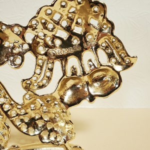Op de afbeelding: Gouden metalen decoratieve standaard met een boomontwerp en de woorden "REVERE MFG" op de achterkant. De standaard heeft een ronde bovenkant en drie poten.