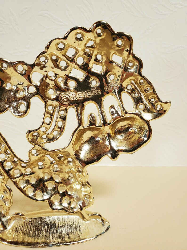 Op de afbeelding: Gouden metalen decoratieve standaard met een boomontwerp en de woorden "REVERE MFG" op de achterkant. De standaard heeft een ronde bovenkant en drie poten.
