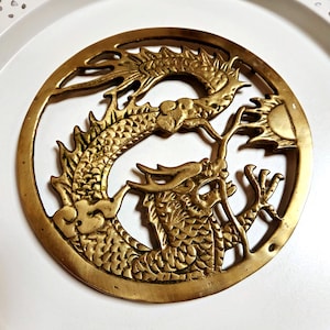 May include: A golden-colored, circular metal dragon sculpture. The dragon is intricately detailed with scales, claws, and a flowing mane. The dragon is positioned in a circular frame, with a sun-like element. The sculpture is likely a decorative item.