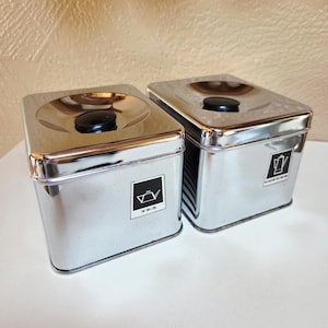 May include: Two shiny, rectangular metal canisters for tea and coffee. Each has a black knob on top and a label with a graphic and text. The tea canister has a teapot graphic and the word "TEA". The coffee canister has a coffee pot graphic and the word "COFFEE".