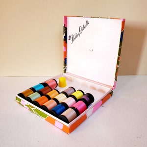May include: A vintage sewing kit with a colorful floral patterned box. Inside, spools of thread in various colors are neatly arranged, alongside a yellow thimble. The box lid is open, displaying the "Belding Corticelli" logo.