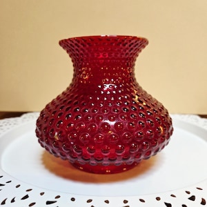May include: A deep red glass vase featuring a distinct hobnail texture across its entire surface. The vase has a wide, rounded body that tapers into a narrower neck and flares slightly at the top opening, creating a classic silhouette.