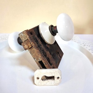 May include: An antique door lock set featuring a heavily rusted metal lock box, two white ceramic door knobs, and a matching keyhole cover. The knobs are oval-shaped and the keyhole cover is rectangular.