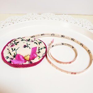 May include: A decorative hat-shaped pin cushion with a floral pattern, pink bow, and gold heart. A beige measuring tape with red markings is coiled nearby. The tape measures up to 63.5 cm.