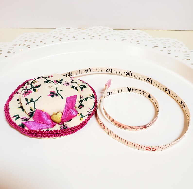 May include: A decorative hat-shaped pin cushion with a floral pattern, pink bow, and gold heart. A beige measuring tape with red markings is coiled nearby. The tape measures up to 63.5 cm.