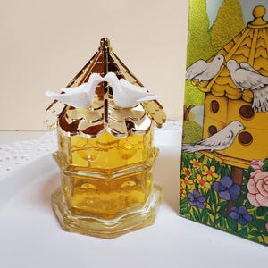 May include: A glass bottle shaped like a birdhouse with a gold roof and two white birds perched on top. The bottle is filled with a clear yellow liquid. The bottle is sitting on a white surface.