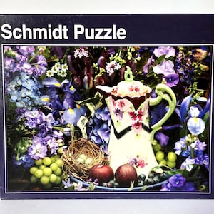 May include: A Schmidt Puzzle box featuring a floral still life. The image includes a decorative teapot with a floral design, surrounded by purple and blue flowers, grapes, and apples. The text "Schmidt Puzzle" is at the top.