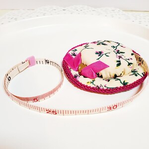 May include: A floral print pin cushion with a pink ribbon and trim, next to a measuring tape. The tape is marked in inches, showing measurements up to 76 cm.