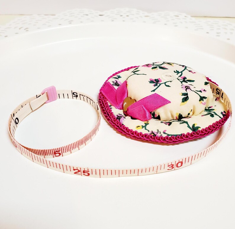 May include: A floral print pin cushion with a pink ribbon and trim, next to a measuring tape. The tape is marked in inches, showing measurements up to 76 cm.