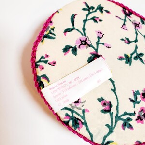 May include: A cream-coloured, heart-shaped item featuring a floral pattern in pink and green. The edge is trimmed with a bright pink, textured border. A white label with text is visible. Made in China for AVON PRODUCTS, INC.