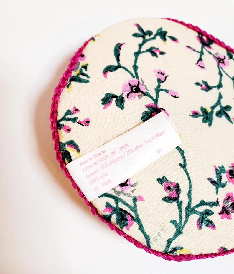 May include: A cream-coloured, heart-shaped item featuring a floral pattern in pink and green. The edge is trimmed with a bright pink, textured border. A white label with text is visible. Made in China for AVON PRODUCTS, INC.