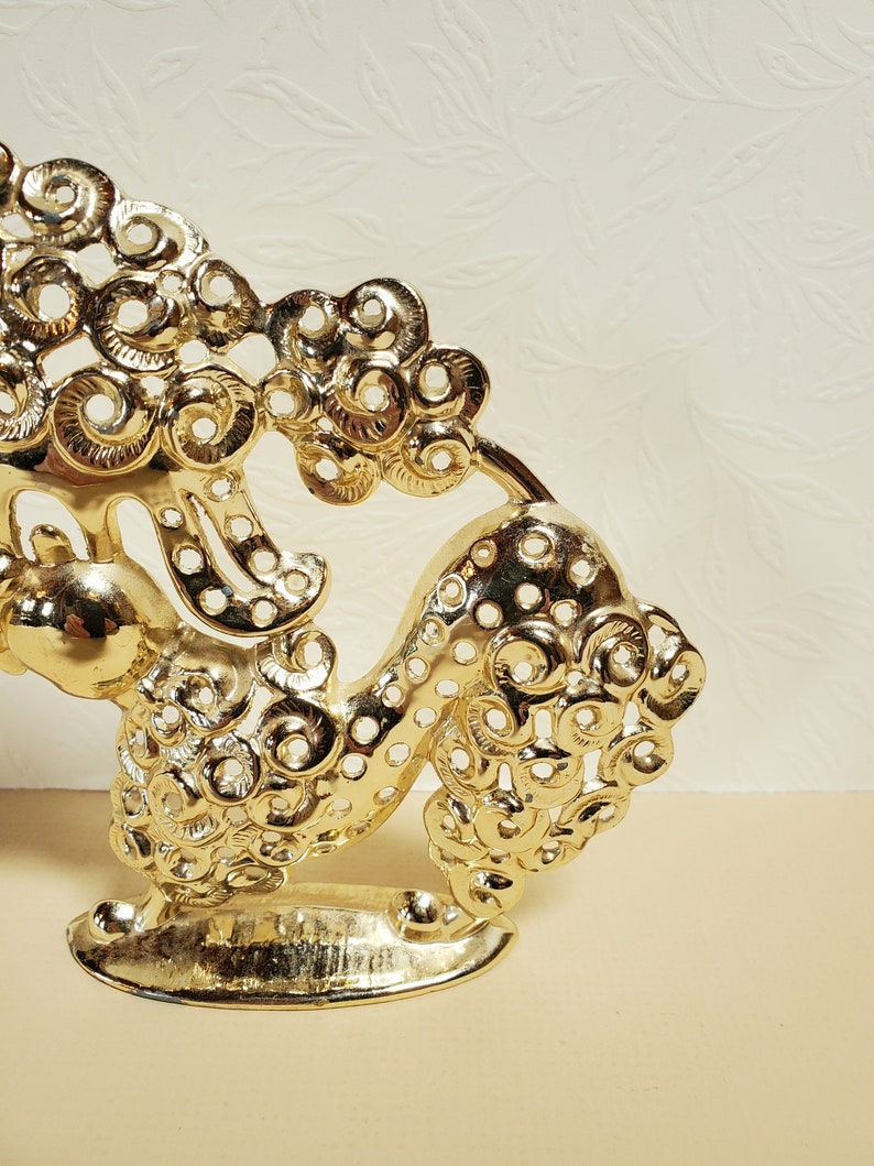 Op de afbeelding: Een gouden metalen decoratief beeldje van een gestileerde draak met ingewikkelde details. De draak staat op een voetstuk en heeft een cirkelvormig ontwerp over zijn hele lichaam.