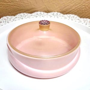 May include: A round, light pink glass powder jar with a smooth, opaque finish. The lid features a small, raised, circular knob at the center, decorated with a cluster of tiny, translucent, purple-tinted glass beads arranged in a floral pattern.