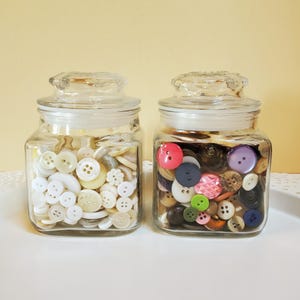 May include: Two glass jars filled with a variety of colorful buttons. The jar on the left contains white and cream buttons, while the jar on the right contains a mix of colors, including pink, purple, green, brown, and blue.