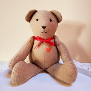 May include: A brown teddy bear with black eyes and a red bow. The bear is sitting on a white surface and has a red heart-shaped button on its chest.