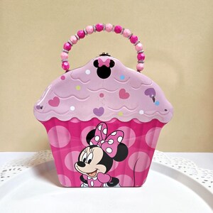 May include: A cupcake-shaped metal tin featuring a pink and white polka dot base with an illustration of Minnie Mouse. The top section is light pink with heart and dot accents and a black Minnie Mouse silhouette. It has a beaded pink handle.