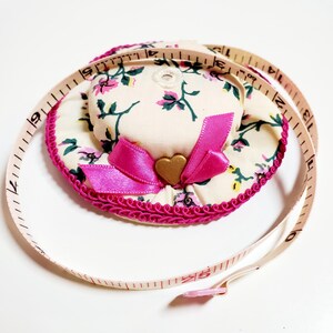 May include: A floral print hat-shaped pin cushion with a pink bow and gold heart accent. The hat is trimmed with pink trim and is next to a white and red measuring tape. The measuring tape is marked in inches and centimeters.
