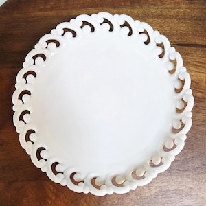 May include: A white, round decorative plate with a scalloped edge. The edge features a repeating pattern of curved cutouts. The plate is made of a glossy material and sits on a wooden surface.