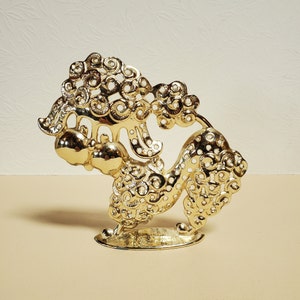 Op de afbeelding: Een gouden metalen sculptuur van een gestileerd dier met ingewikkelde ronde patronen. Het beeld heeft een rond hoofd en een lange, gekrulde staart.