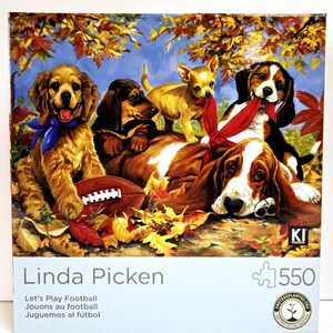 May include: A 550-piece jigsaw puzzle featuring a painting by Linda Picken. The artwork depicts five dogs of different breeds, including a golden retriever, dachshund, chihuahua, and beagle, with a football and autumn leaves. The puzzle's title is "Let's Play Football."